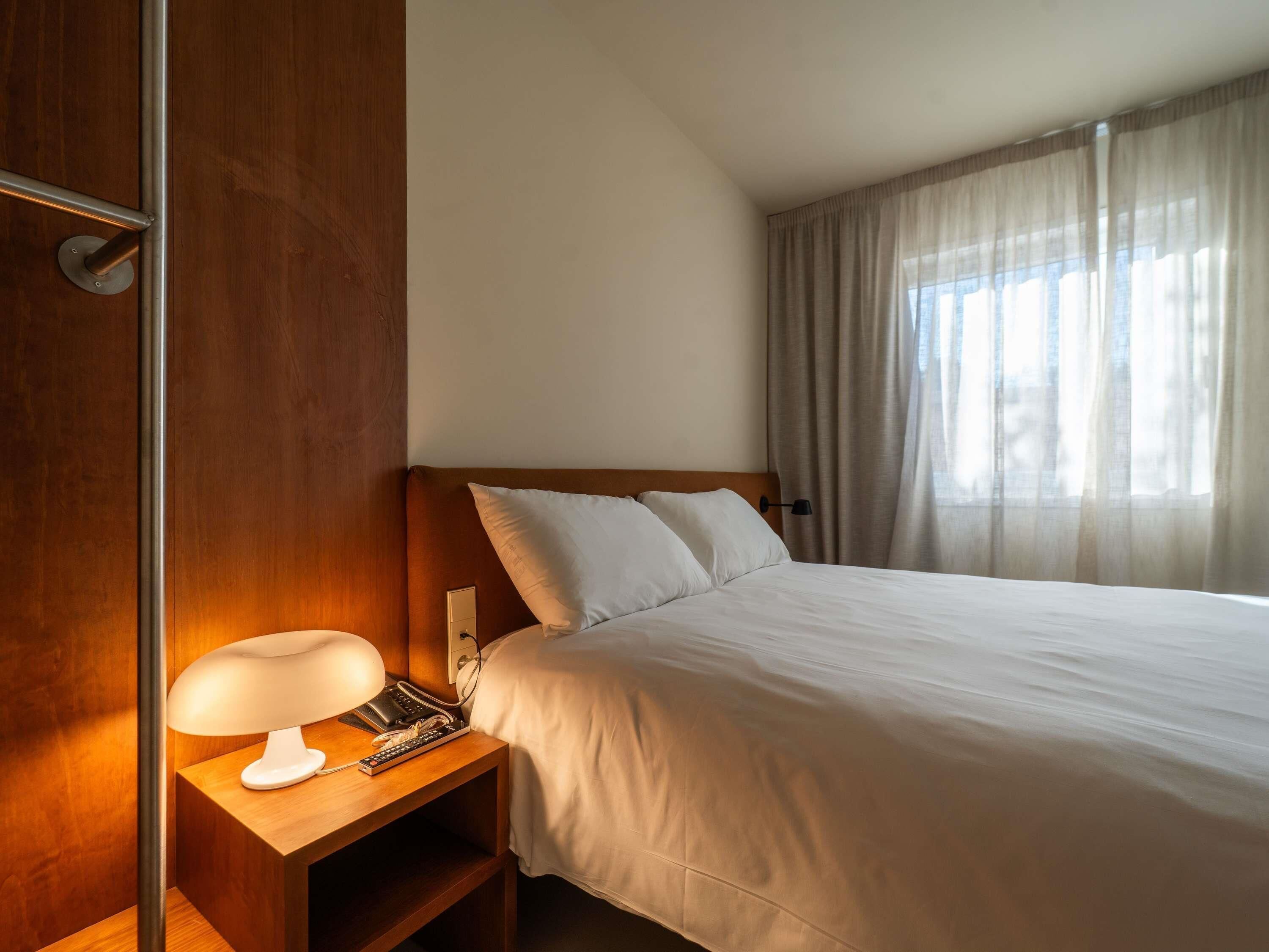 A hotel room with a bed, a bedside lamp, a telephone, and a window with curtains.