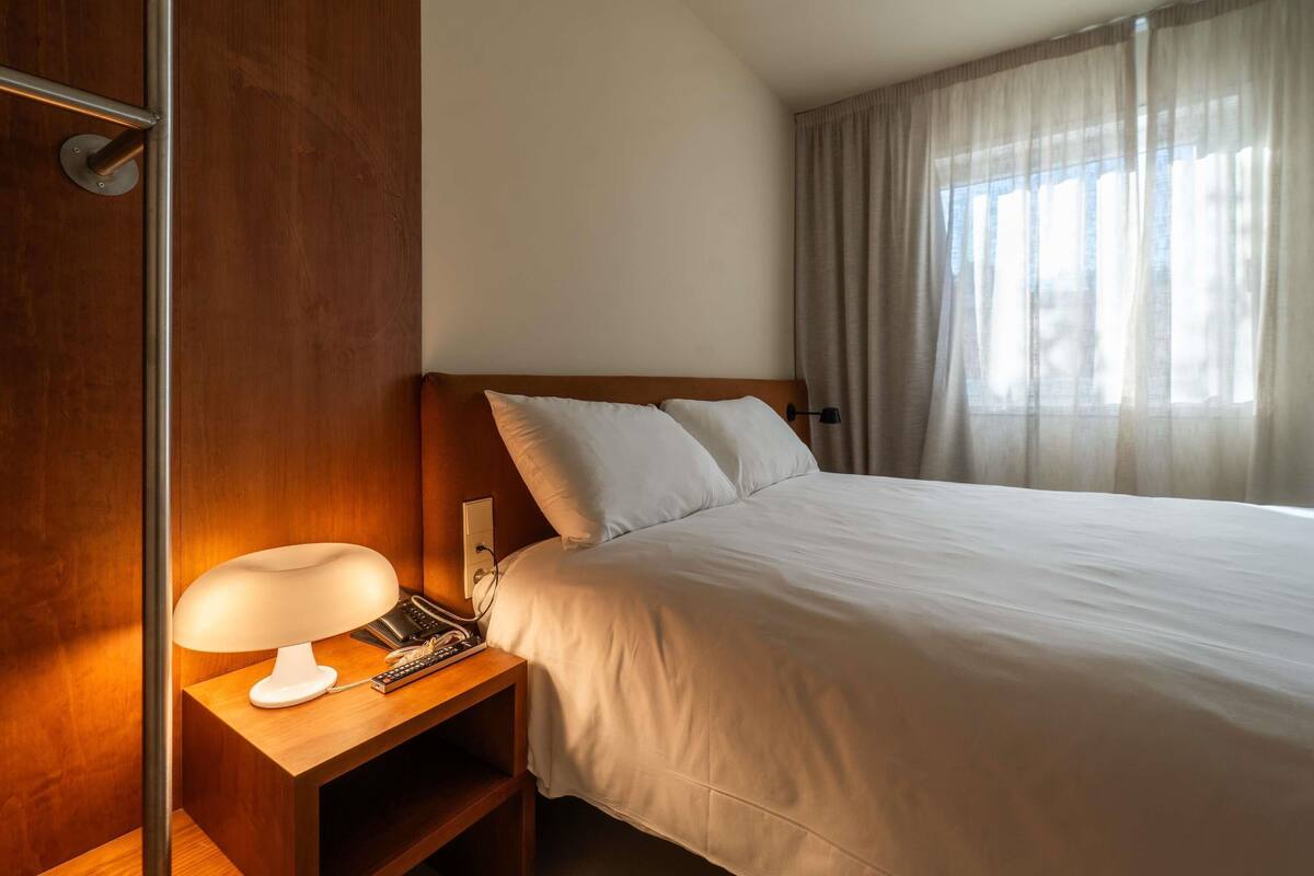 A hotel room with a bed, a bedside lamp, a telephone, and a window with curtains.