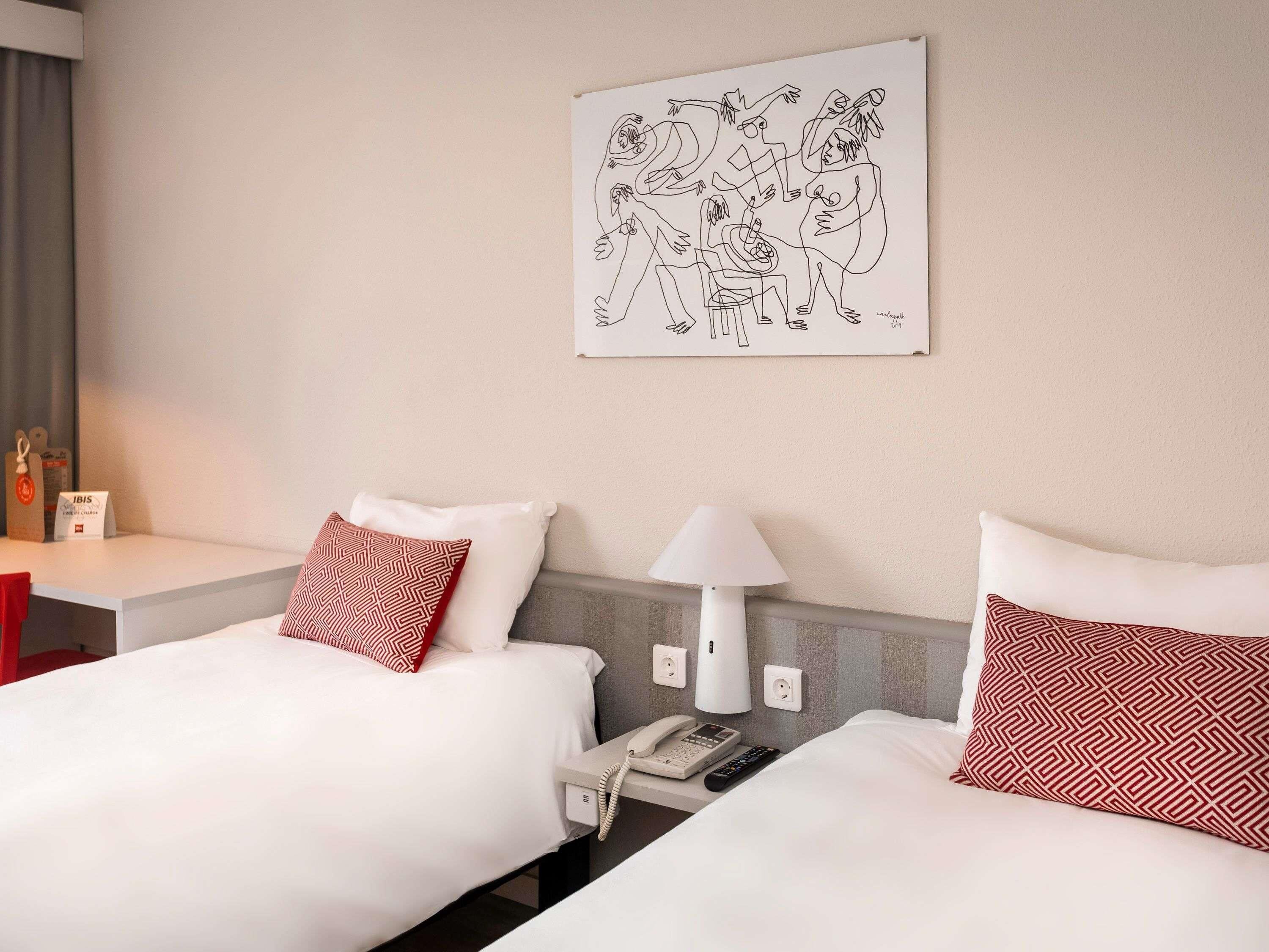 A hotel room with two beds, a bedside table with a lamp, a phone, and a wall-mounted artwork.