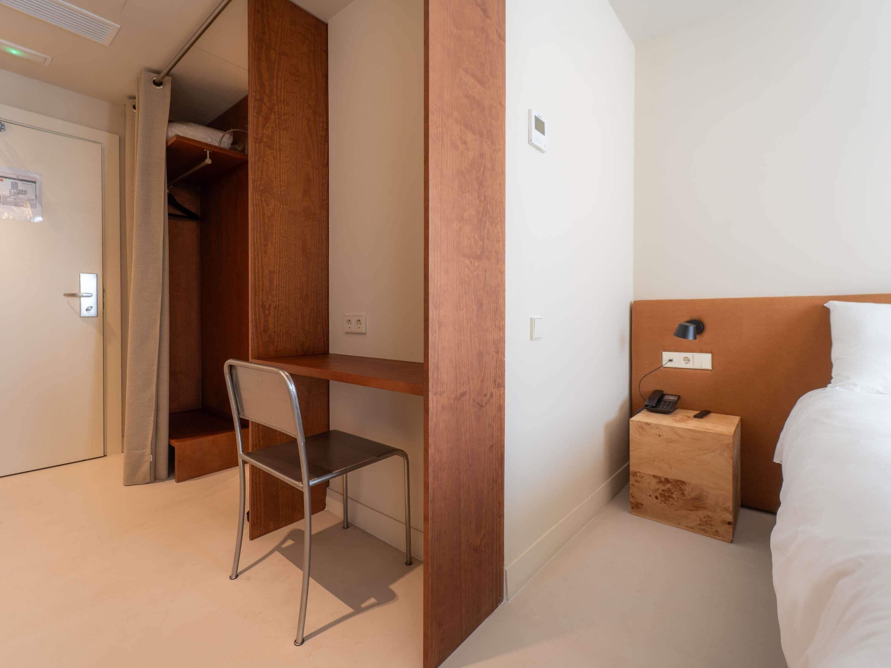 A hotel room with a bed, a desk, a chair, a wardrobe, and a bedside table with a lamp.