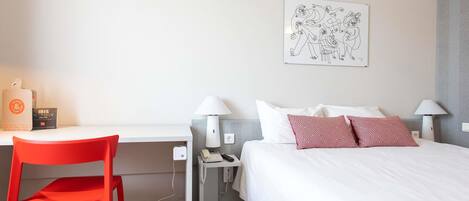 A hotel room with a bed, a desk, a chair, a lamp, a phone, and a wall art piece.