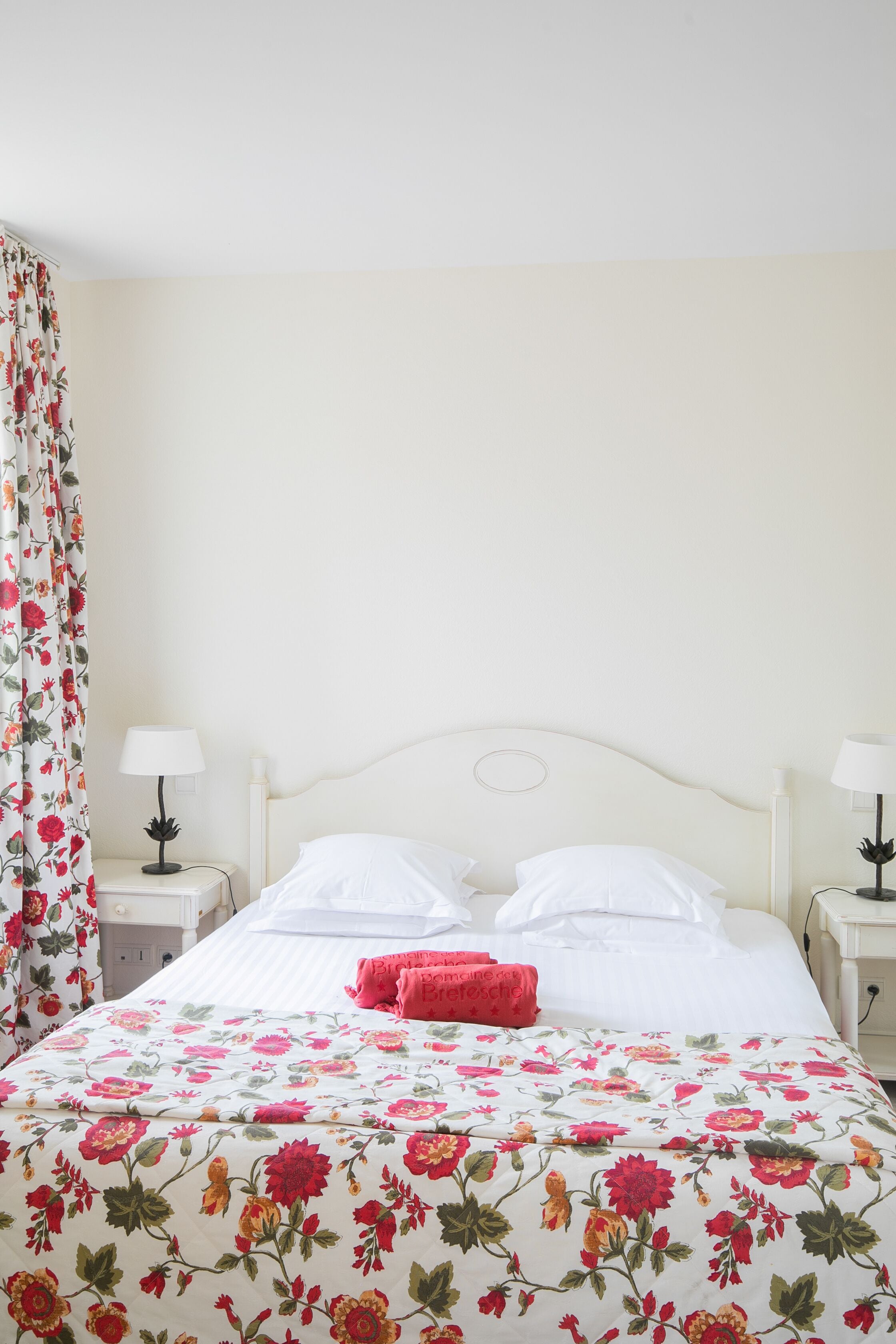 Un lit bien fait, recouvert d’une couvre-lit à motifs floraux, deux tables de chevet avec des lampes et un rideau à fleurs.