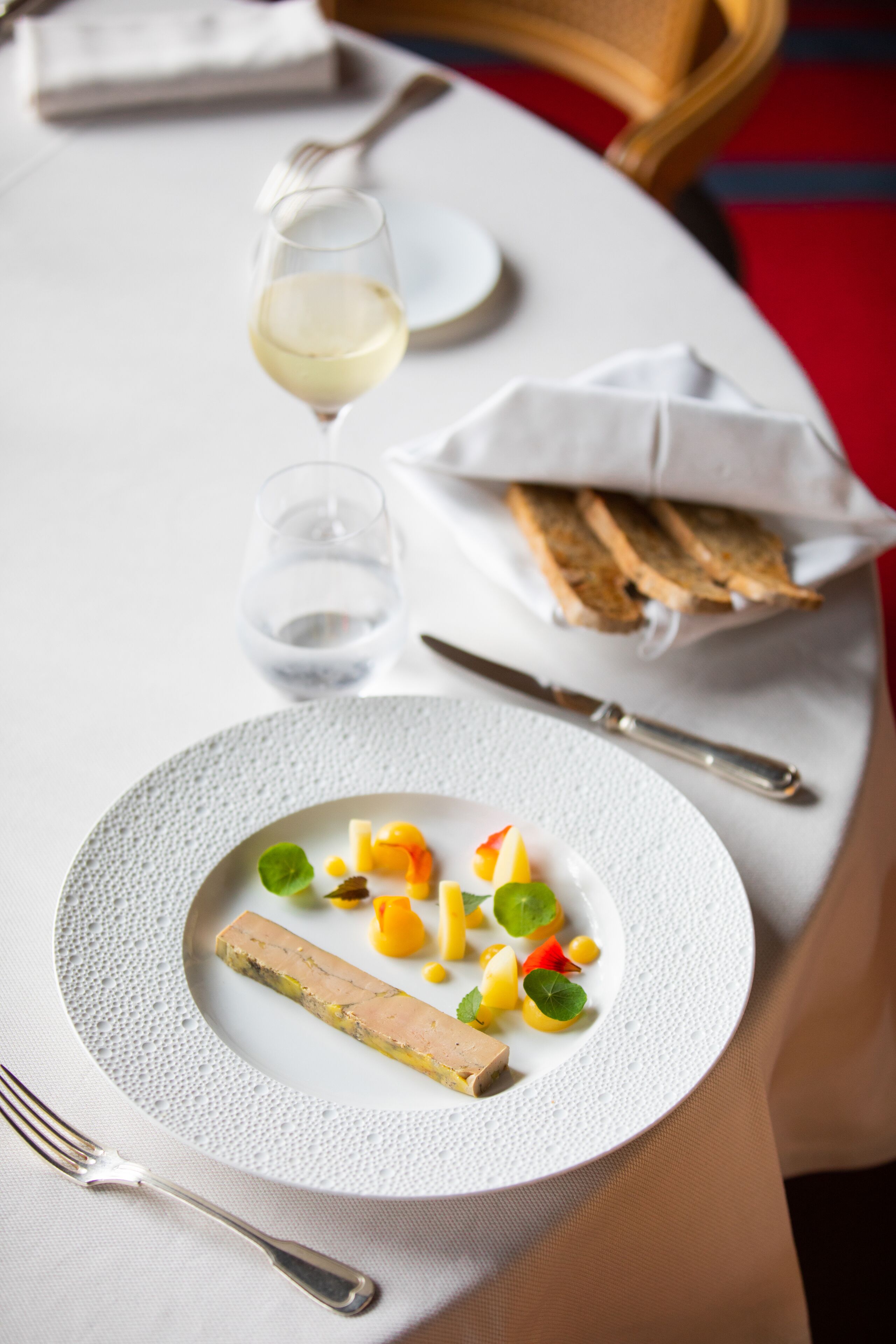 Une table à manger avec une assiette de nourriture, une fourchette, un couteau et un verre de vin blanc.