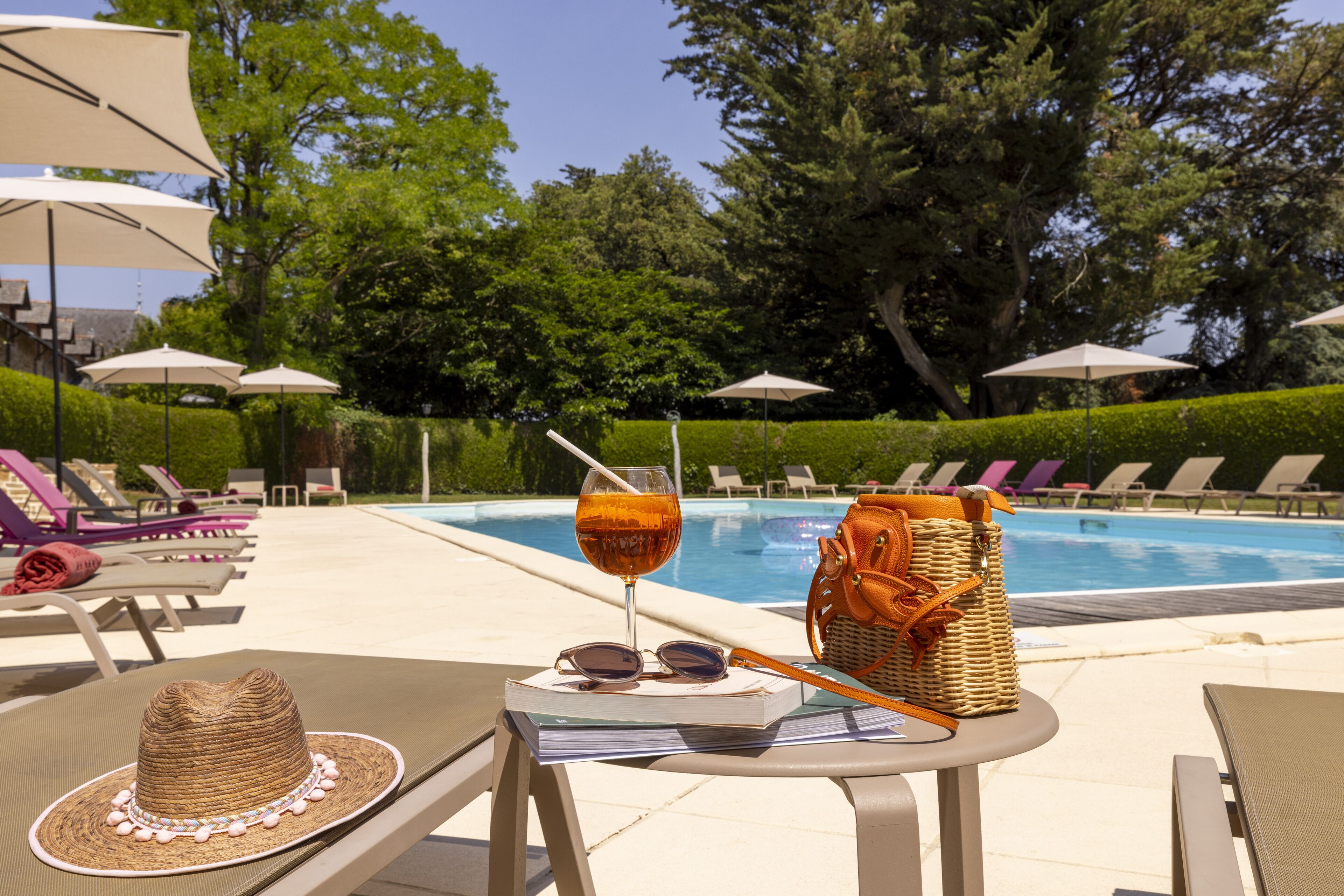 A poolside setting with lounge chairs, umbrellas, a wicker basket, a book, a straw, and a drink.