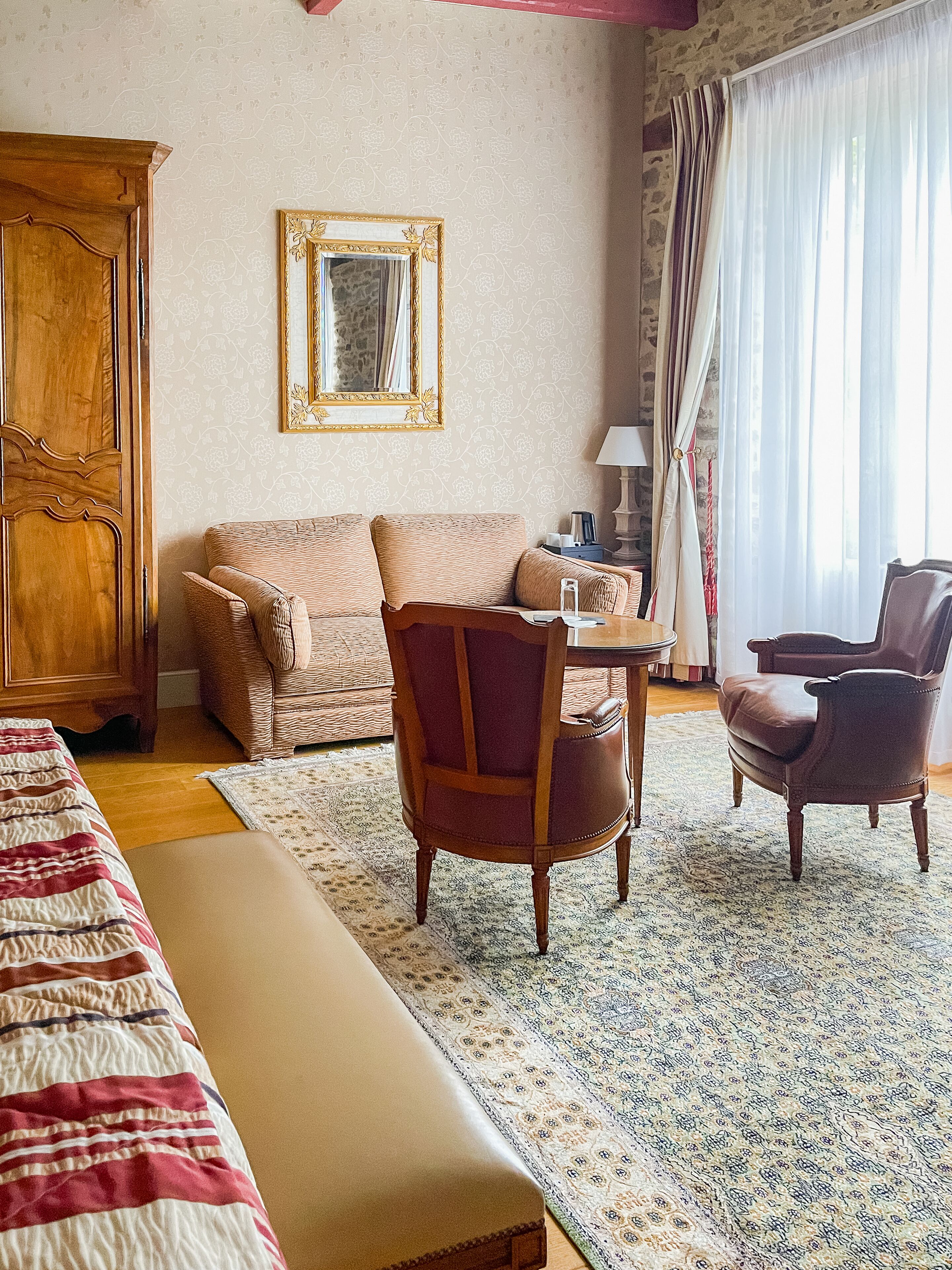 A hotel room with a bed, a sofa, two armchairs, a wooden cabinet, a mirror, and a window with curtains.