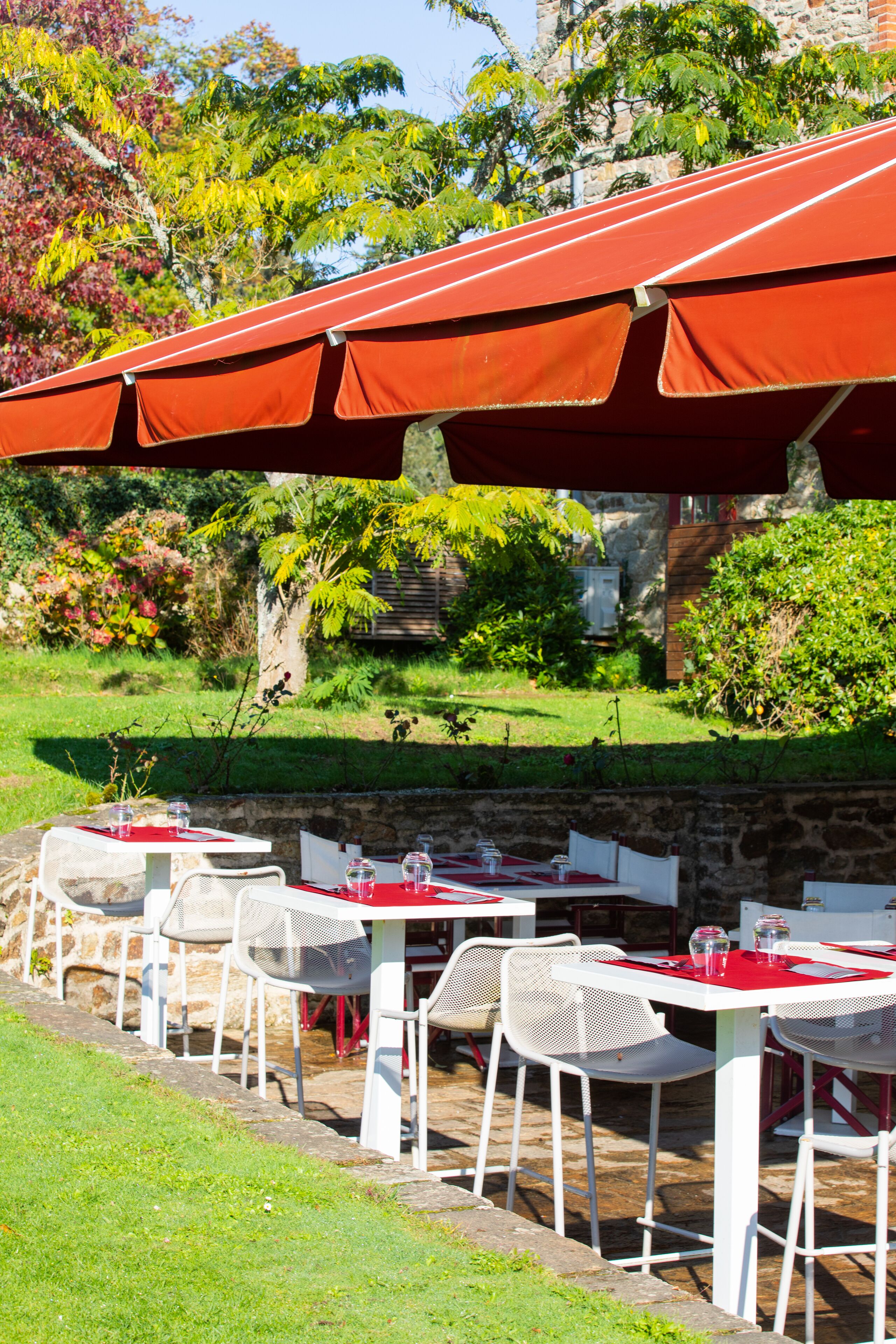 Un restaurante al aire libre con mesas blancas y sombrillas rojas, rodeado de follaje y muros de piedra.