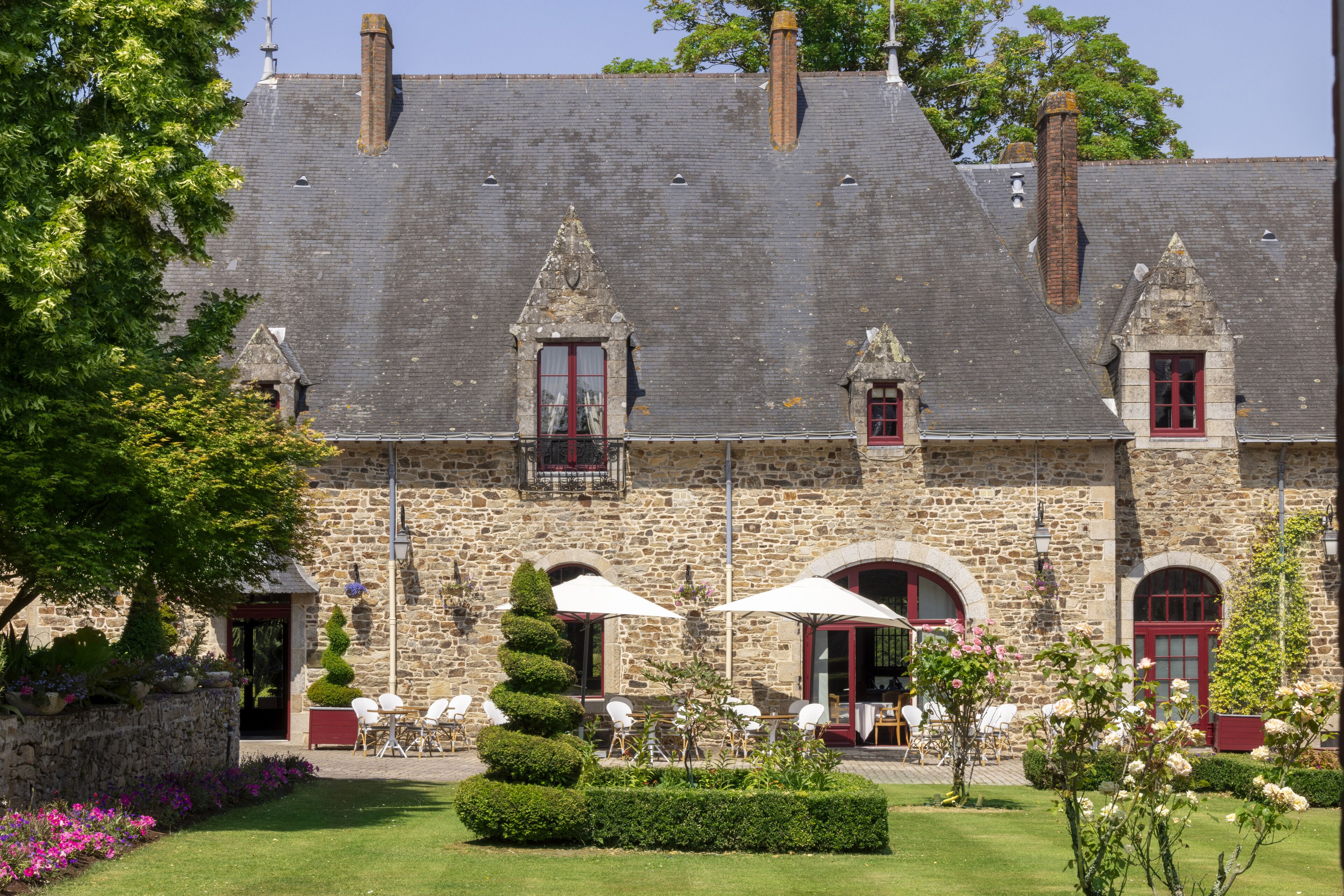 A stone building with a sloped roof, multiple chimneys, and red-framed windows. There are outdoor seating areas with white umbrellas and chairs.
