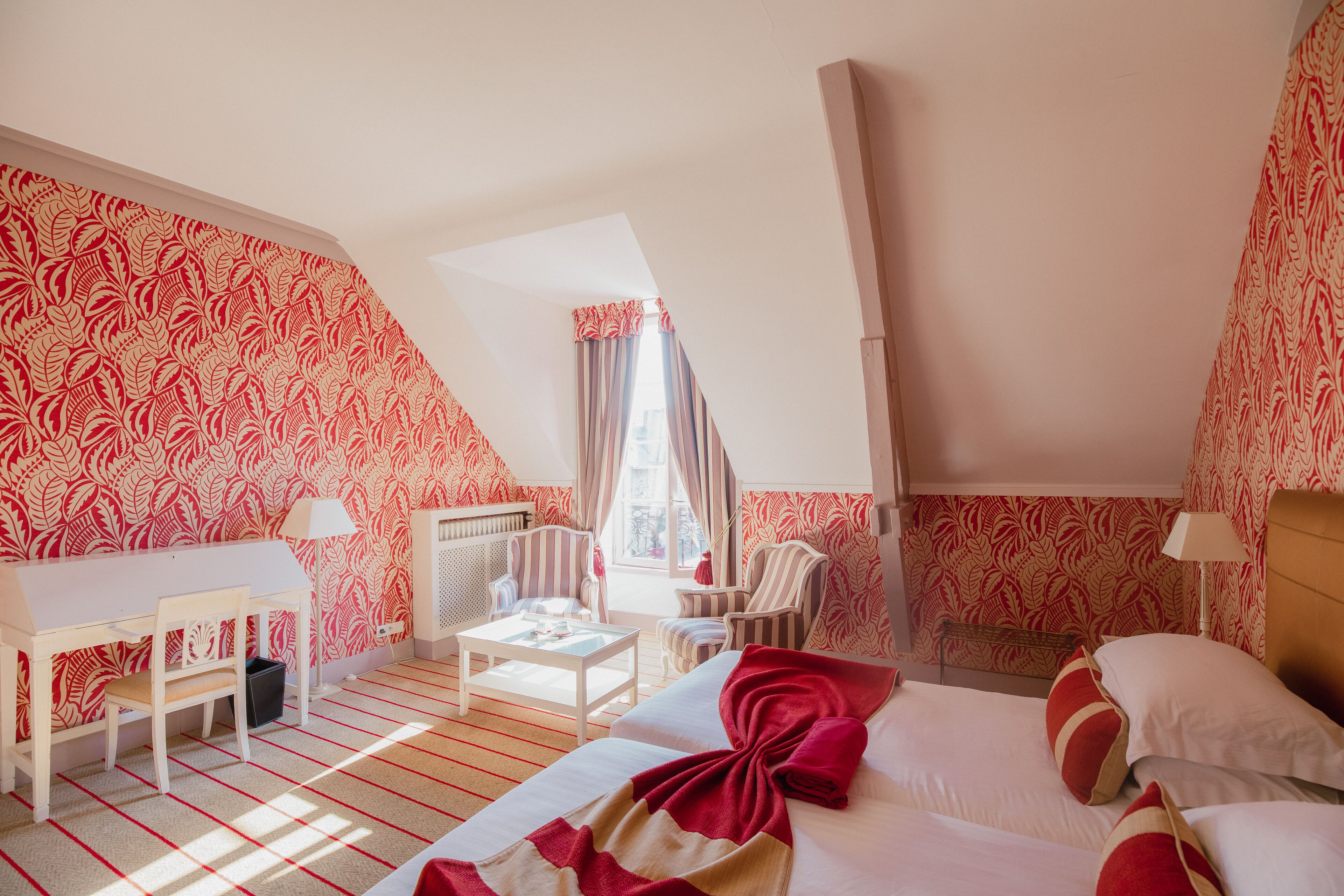 A hotel room with a sloped ceiling, a bed with red and white bedding, a desk with a chair, a small table, and a radiator.