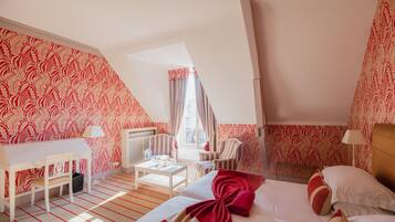 A hotel room with a sloped ceiling, a bed with red and white bedding, a desk with a chair, a small table, and a radiator.