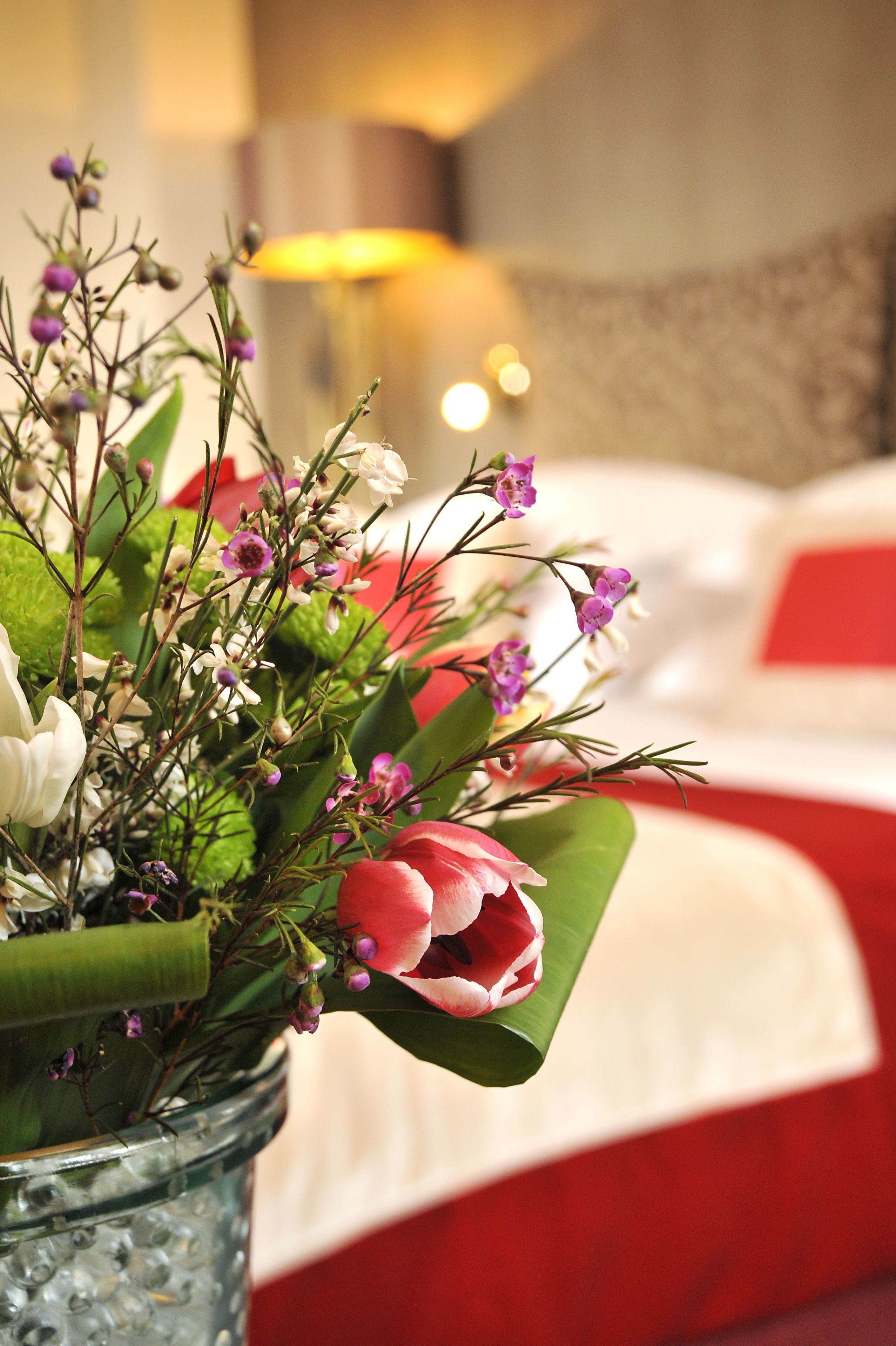 A bouquet of flowers in a glass vase on a bed with red bedding.