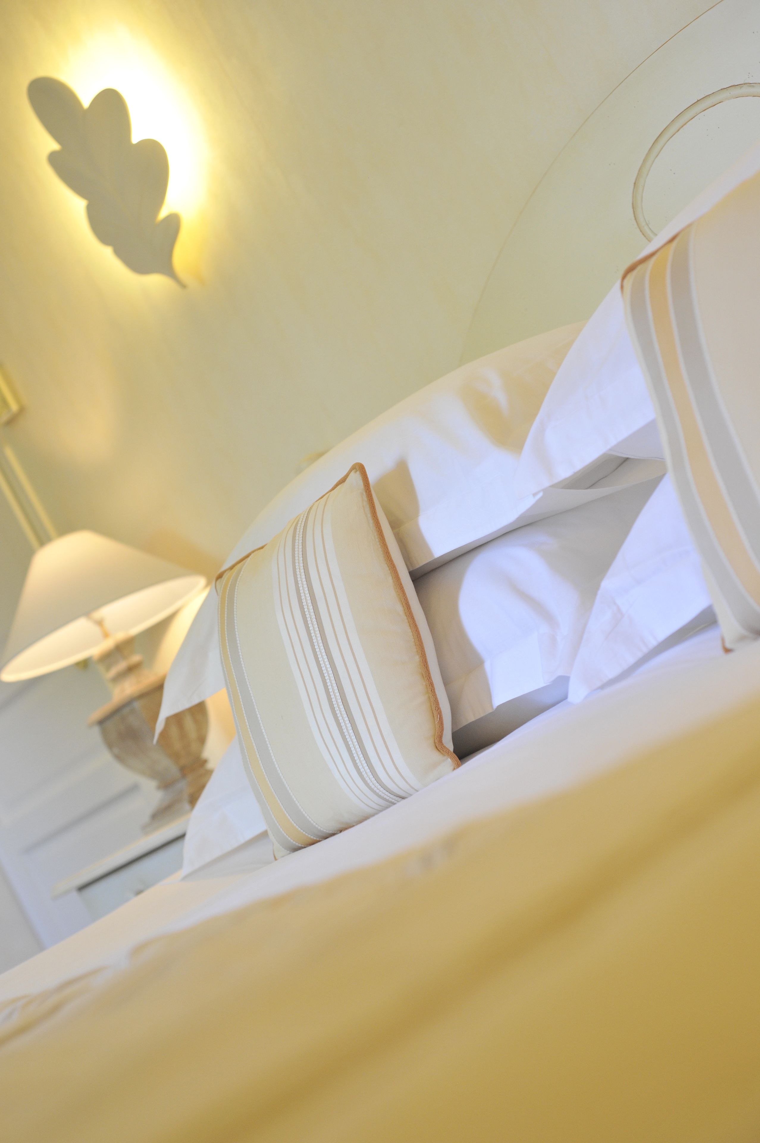 Un lit bien fait, avec du linge de lit blanc et des oreillers, un luminaire fixé au mur et une table de chevet avec une lampe.