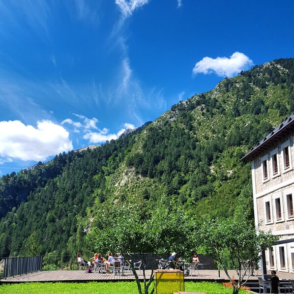Una terraza con mobiliario exterior, un edificio de piedra y una montaña boscosa al fondo.