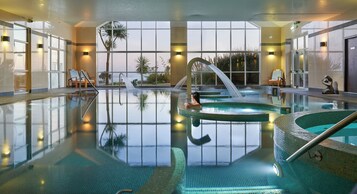 A large indoor swimming pool with clear glass walls, multiple seating areas, and a view of palm trees outside.