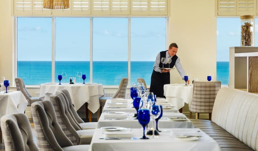 A restaurant with a view of the ocean, tables set with blue glassware, and a waiter attending to guests.