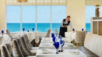 A restaurant with a view of the ocean, tables set with blue glassware, and a waiter attending to guests.