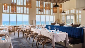 Un restaurant où les tables sont dressées pour le service, avec de grandes fenêtres donnant sur la mer et une vue imprenable sur l’océan.
