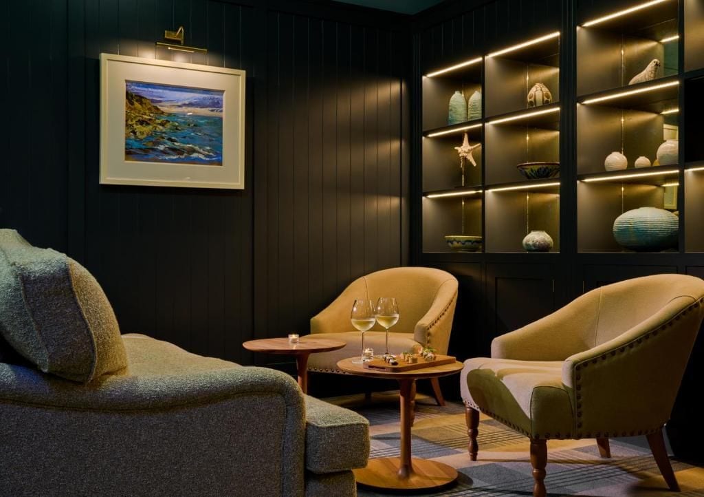 A room with dark wood paneling, a framed painting, a shelf with decorative items, and two armchairs with a small table between them.