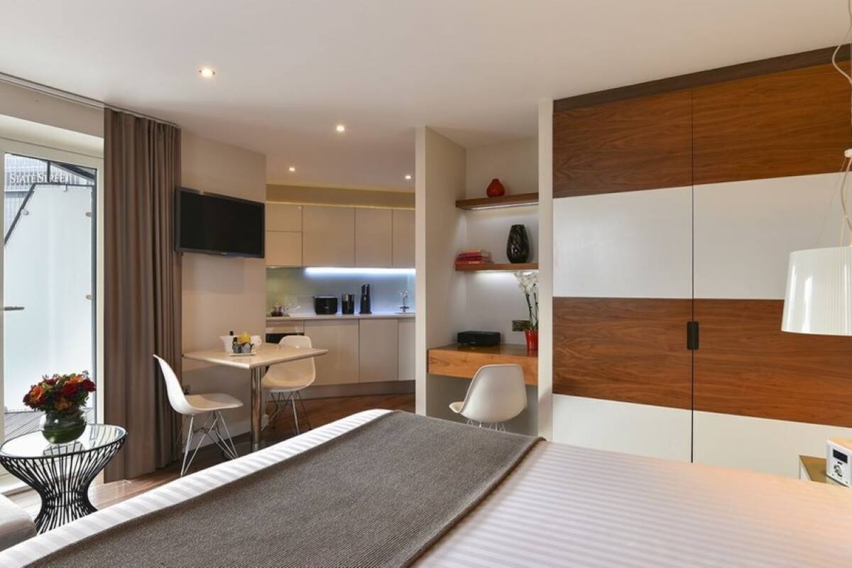 A modern hotel room with a bed, a dining area, and a kitchenette.
