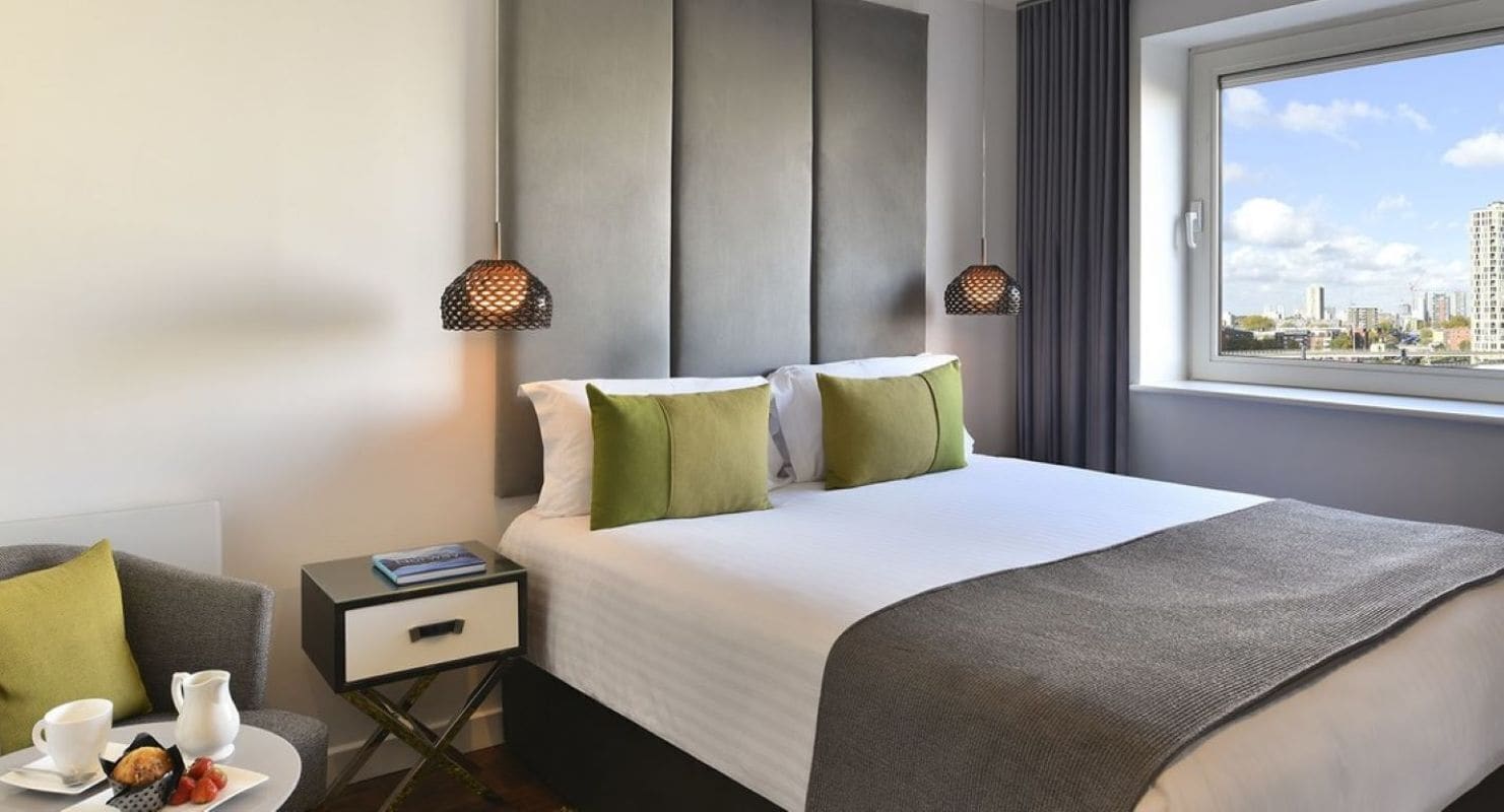 Une chambre d’hôtel moderne, équipée d’un lit, d’une table de chevet et d’une fenêtre donnant sur la ville.
