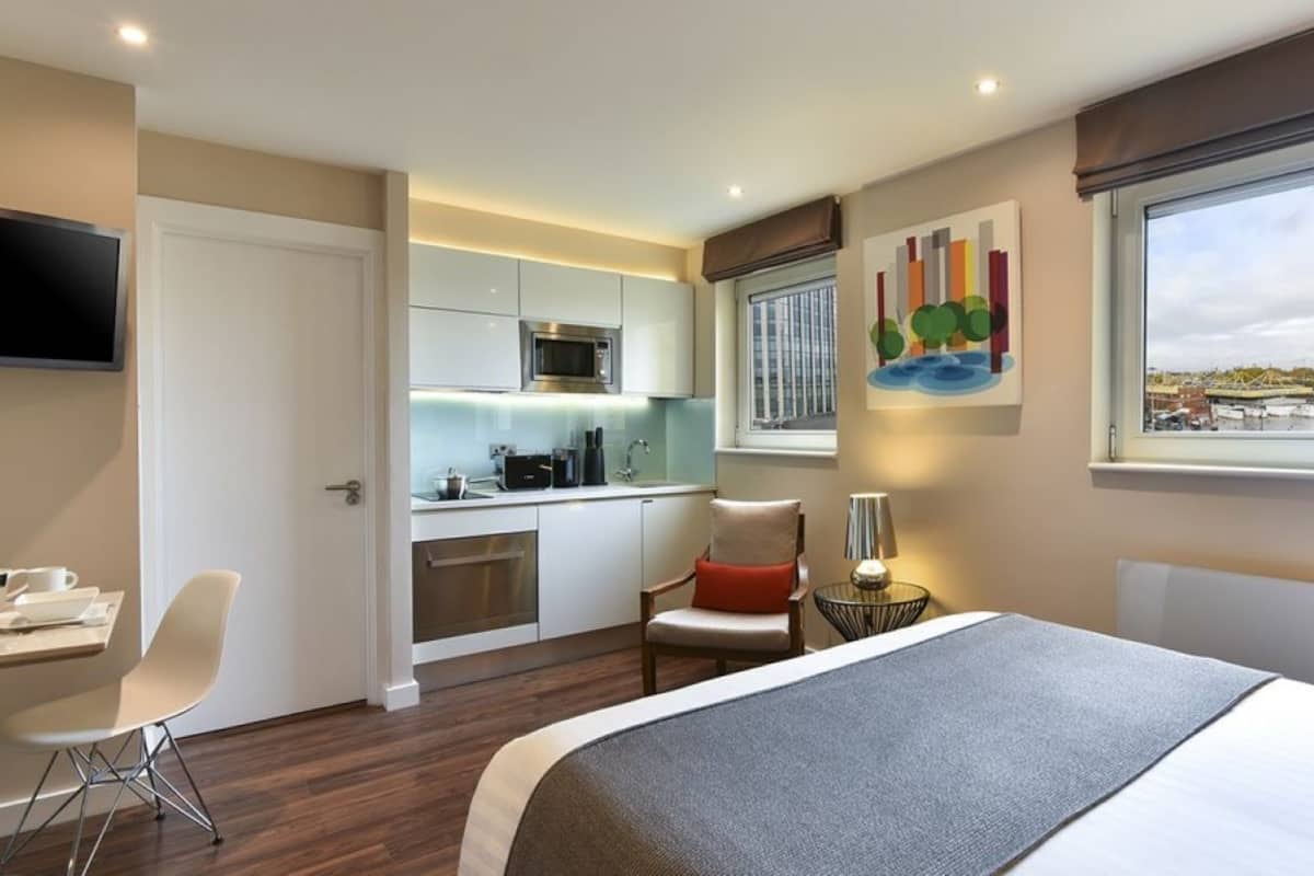 A hotel room with a bed, a desk, a chair, a small table with a lamp, a microwave, and a painting on the wall.
