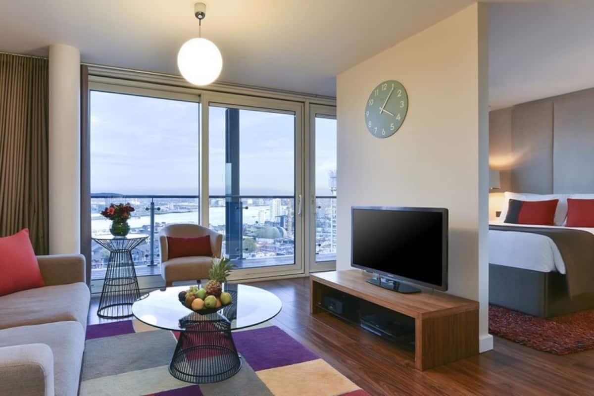 A modern hotel room with a large window, a flat-screen TV, a sofa, a coffee table with fruit, and a bed with red and white bedding.