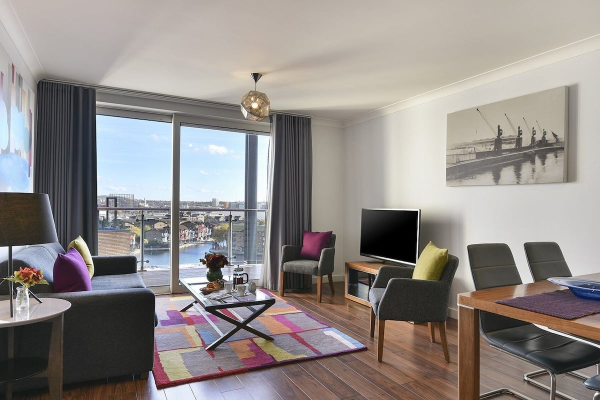 A modern living room with a large window, a flat-screen TV, a dining area, and a view of a cityscape.
