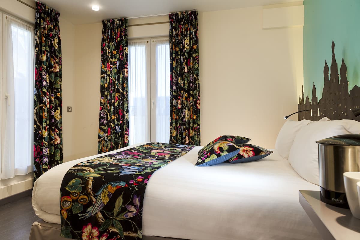 Habitación de hotel con una cama grande, cortinas florales, un mural con vista a la ciudad y una cafetera.