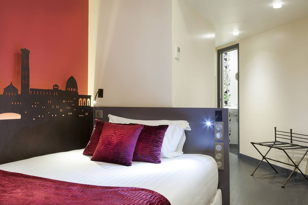 Habitación de hotel con una cama bien hecha, un mural de la ciudad en la pared y un enchufe visible en la mesita de noche.