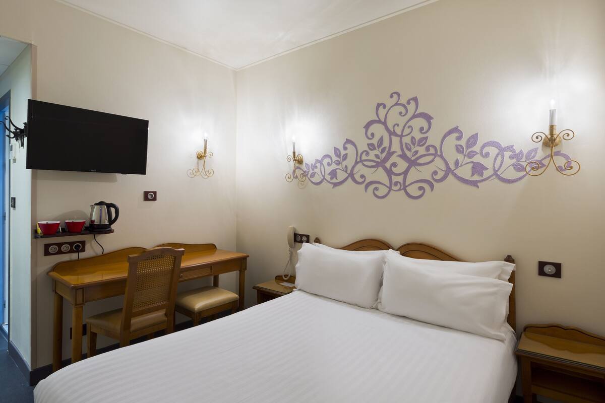 Habitación de hotel con cama, escritorio con silla, televisor montado en la pared y un elemento decorativo en la pared.