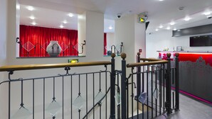 Un vestíbulo de hotel con un fondo de cortina roja, una mesa de recepción y una escalera con barandilla metálica.