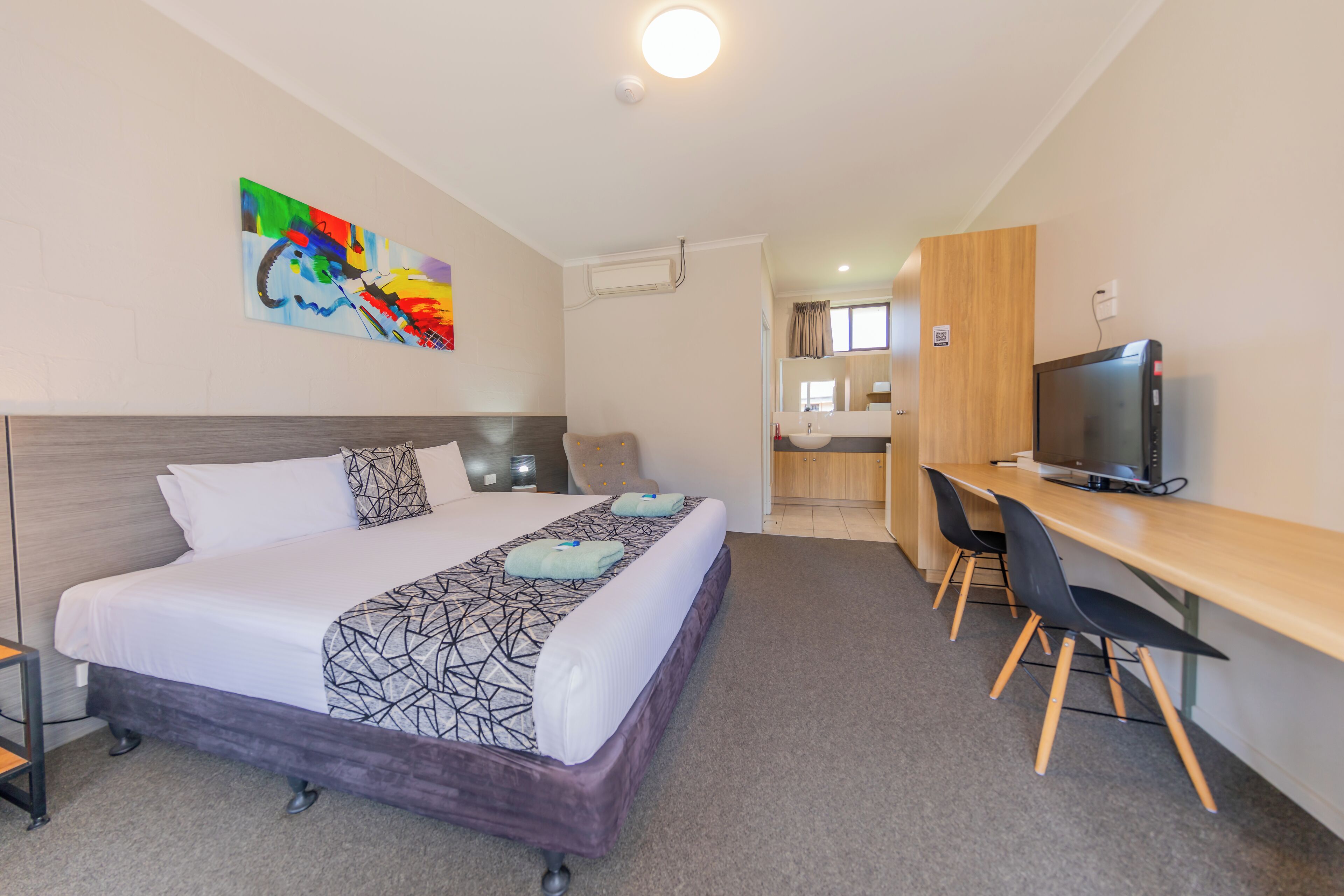 A hotel room with a bed, a desk, a TV, and a bathroom visible in the background.
