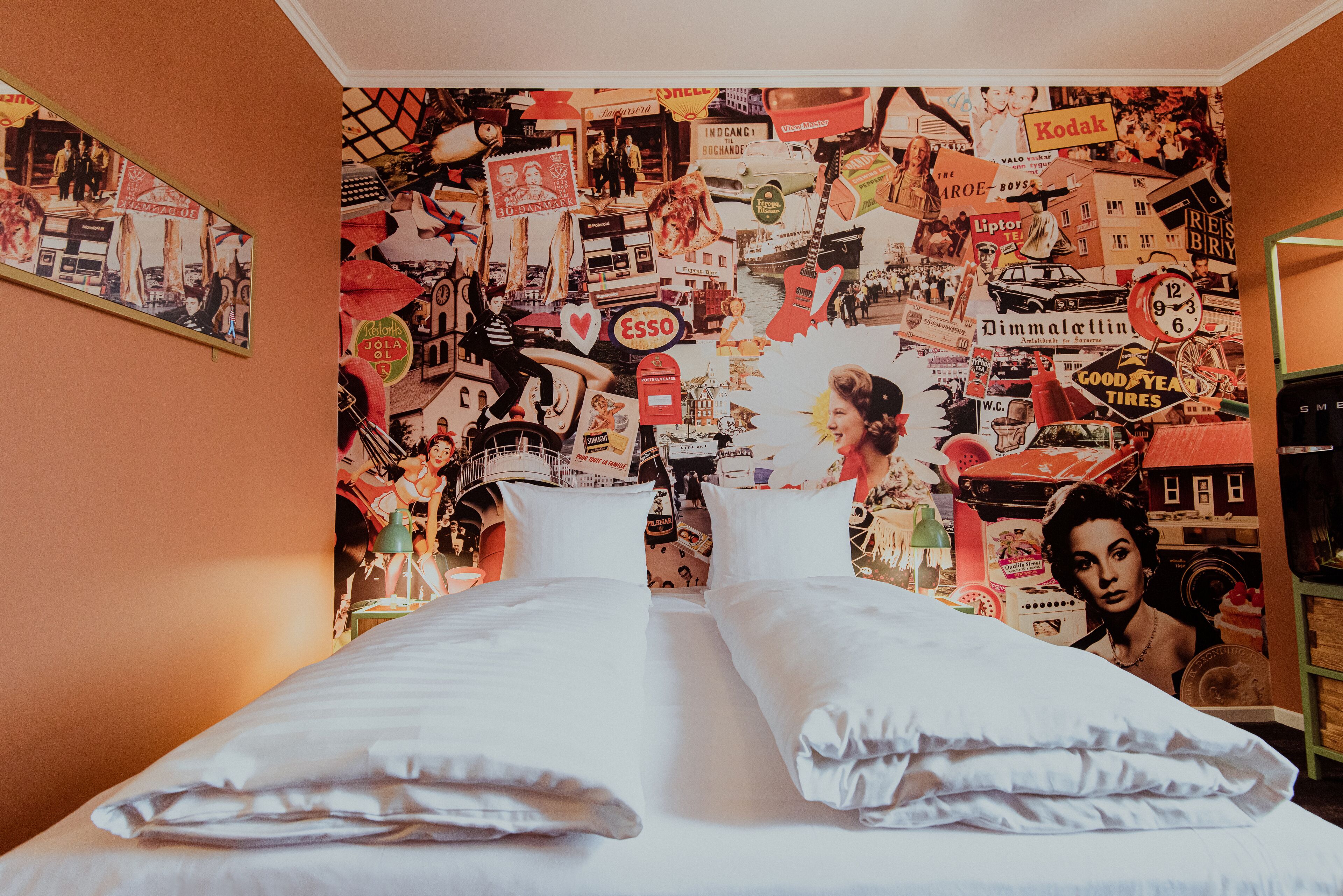 A bed with white linens in a room with a wall covered in various vintage advertisements and images.