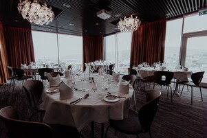 Un restaurante con mesas redondas dispuestas para comer, grandes ventanales que ofrecen vistas a la ciudad y candelabros modernos.