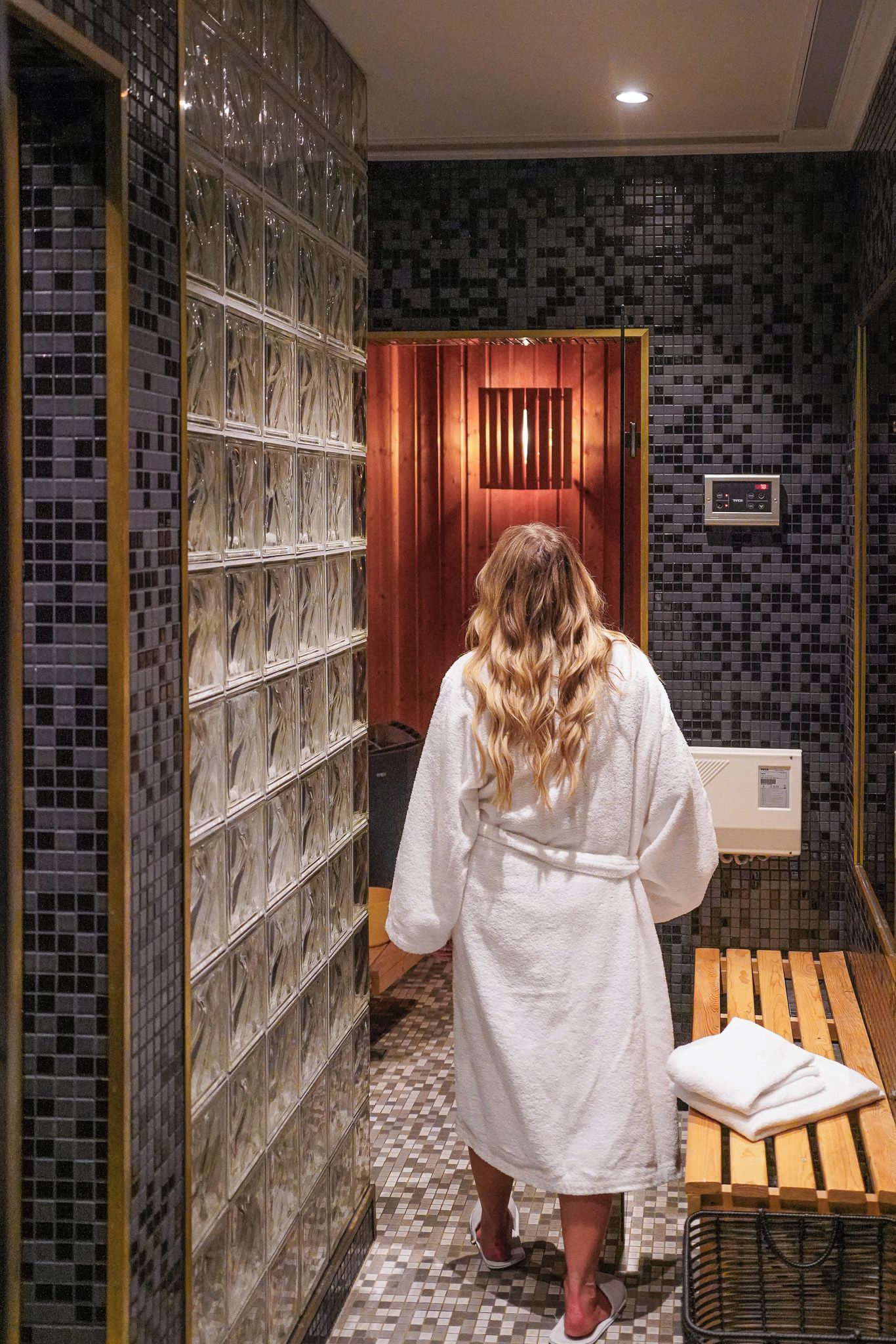 A person in a white robe walking through a hallway with tiled walls and a wooden door.