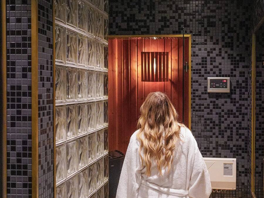 A person in a white robe walking through a hallway with tiled walls and a wooden door.