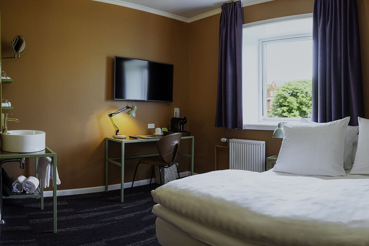 Une chambre d’hôtel comprenant un lit, un bureau avec un ordinateur, une télévision, une fenêtre avec des rideaux et un radiateur.