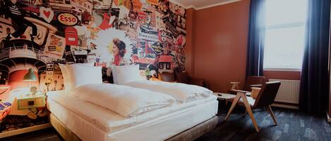 A hotel room with a large bed, a desk, and a wall covered in various posters and advertisements.