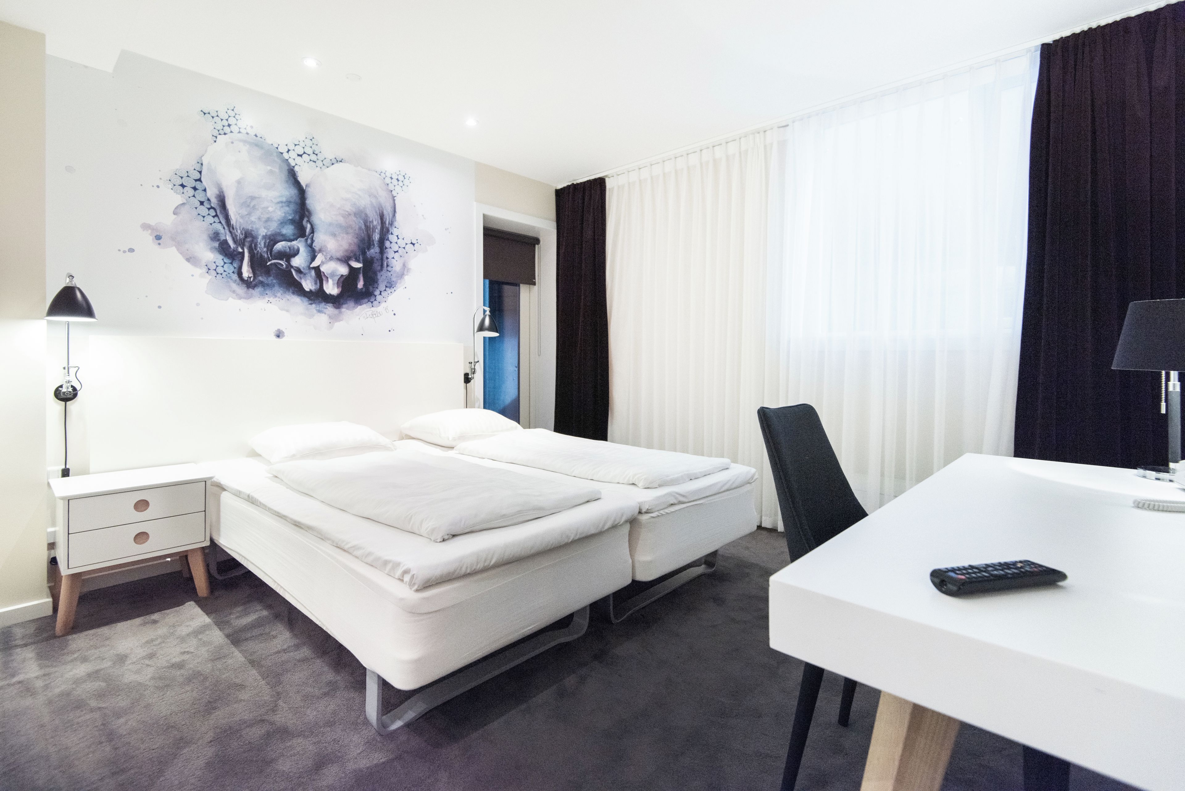 Habitación de hotel con cama, escritorio, silla, lámpara y un cuadro en la pared.