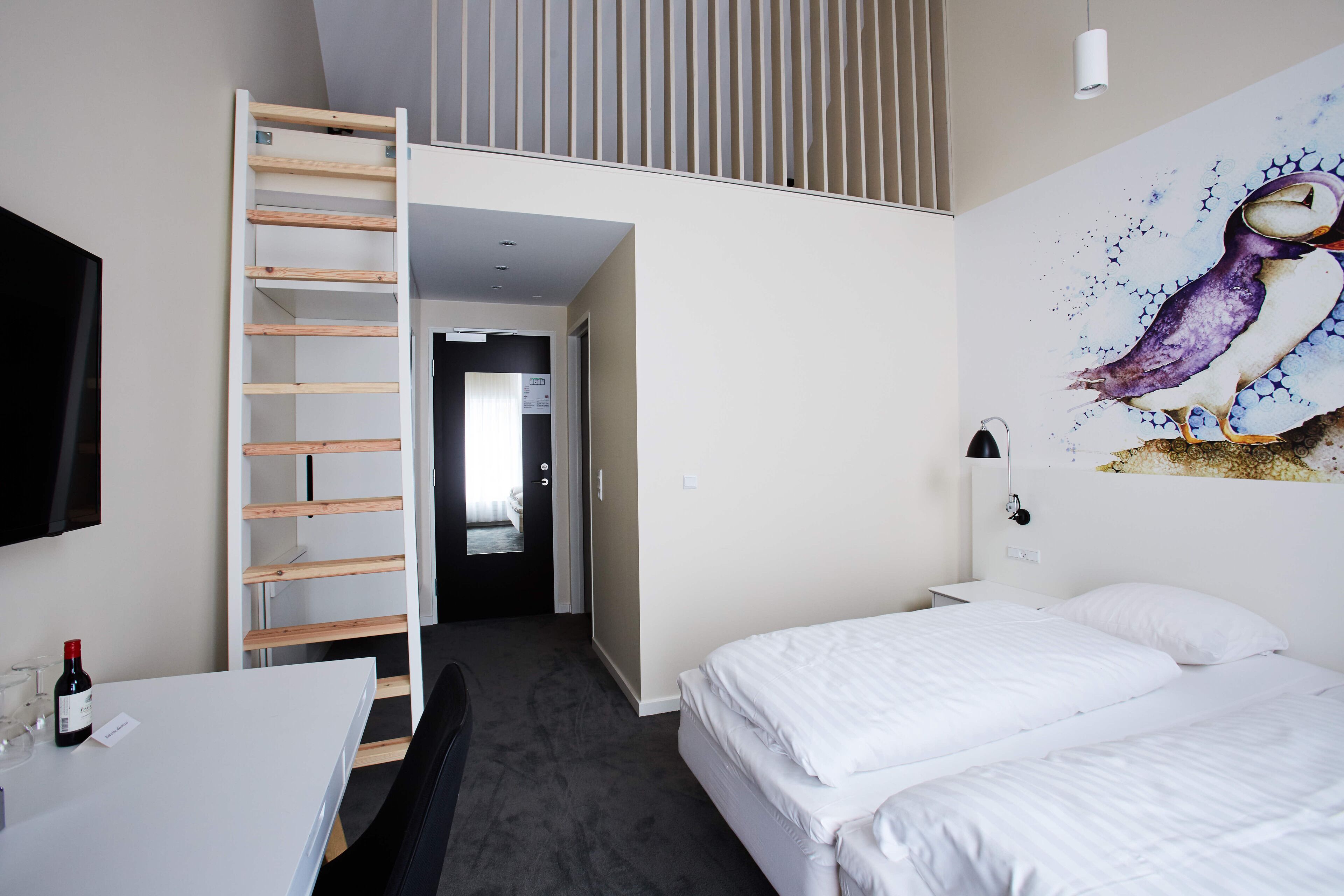 A bedroom with a loft bed, a desk with a wine bottle, and a wall-mounted TV.