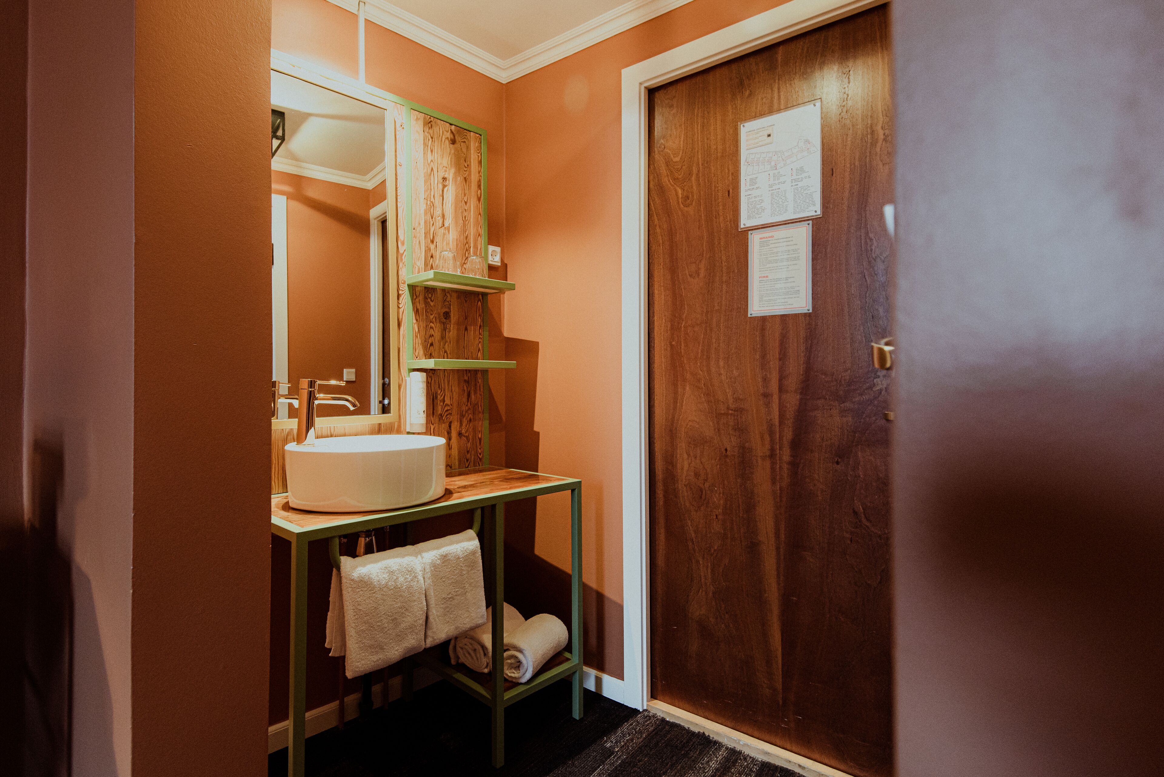 Un baño con puerta de madera, lavamanos con espejo y un perchero para toallas.