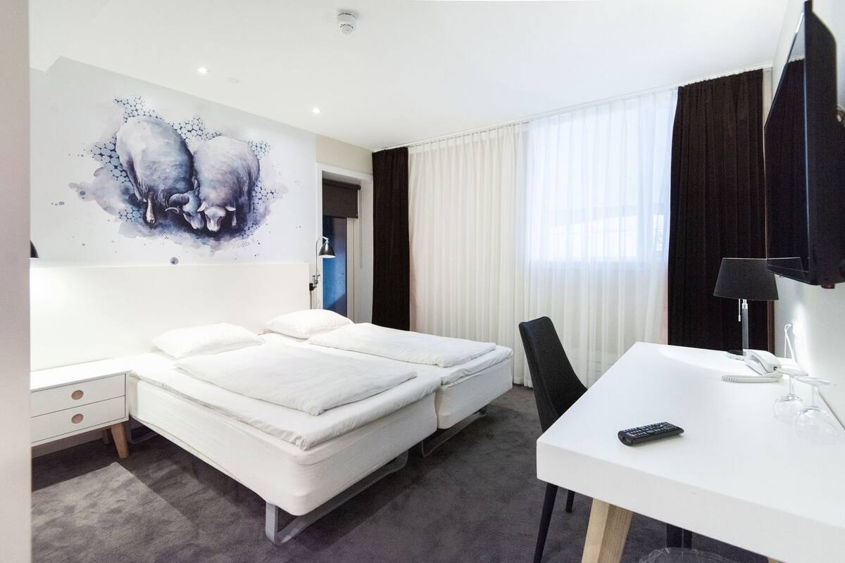 Une chambre d’hôtel avec un lit, un bureau, une chaise, une télévision et un grand tableau abstrait accroché au mur.