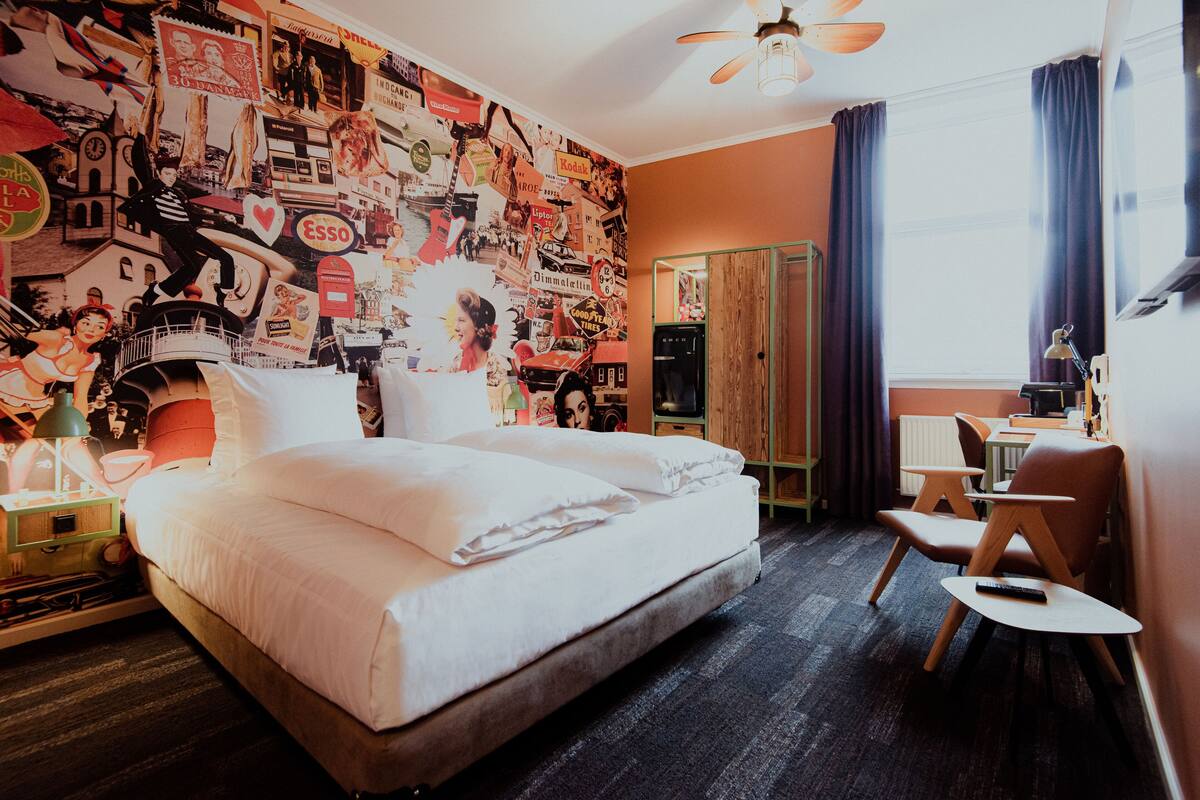 Une chambre d’hôtel avec un grand lit, un bureau et une chaise. La chambre est dotée d’un mur orné de divers affiches et d’une fenêtre avec des rideaux.