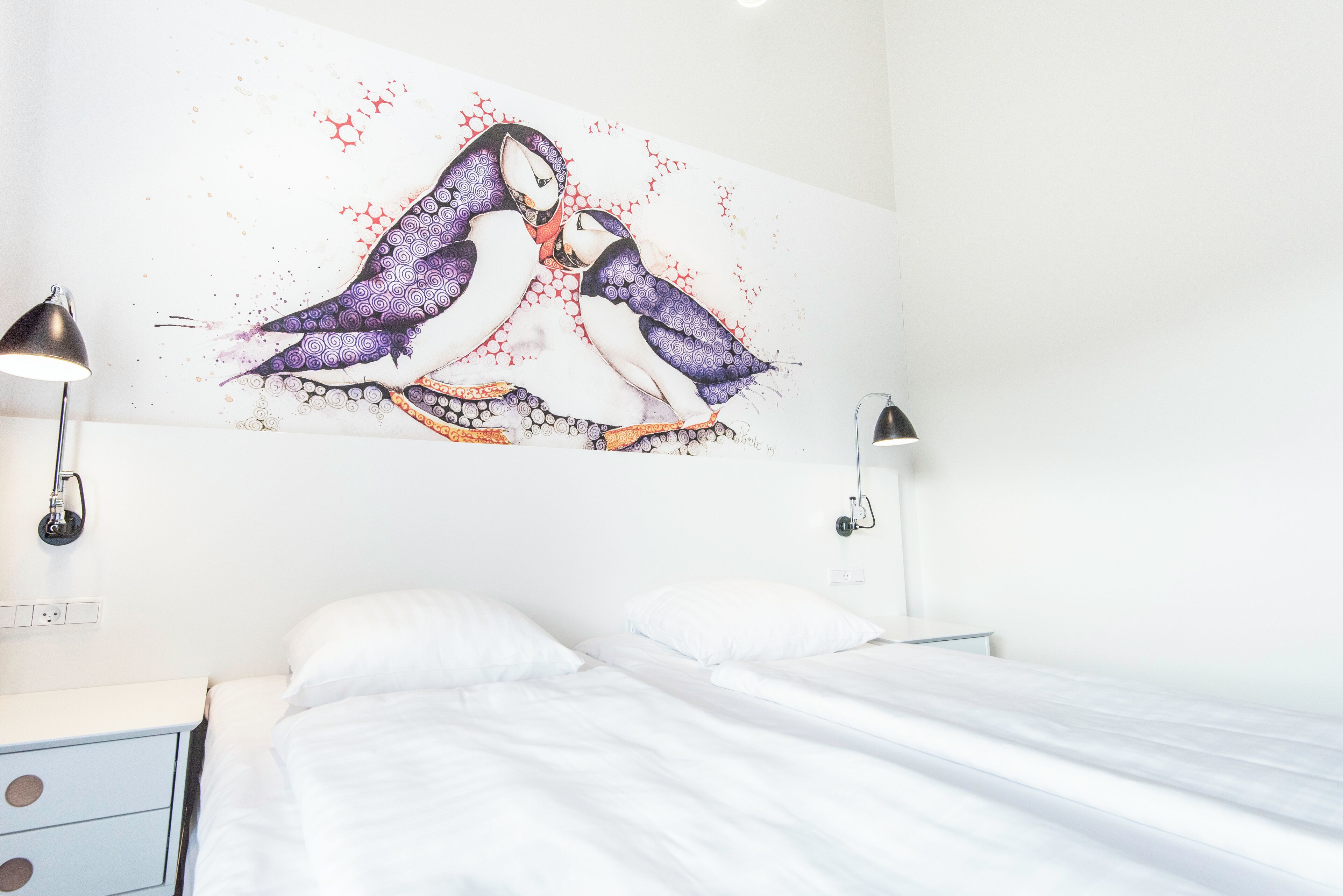 Une chambre d’hôtel avec un lit, des murs blancs et une œuvre d’art représentant deux pingouins, accrochée au mur.