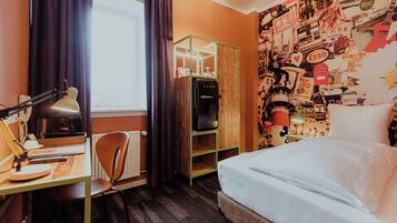 Une pièce comprenant un lit, un bureau avec une lampe, un radiateur et un mur orné de divers affiches et tableaux.