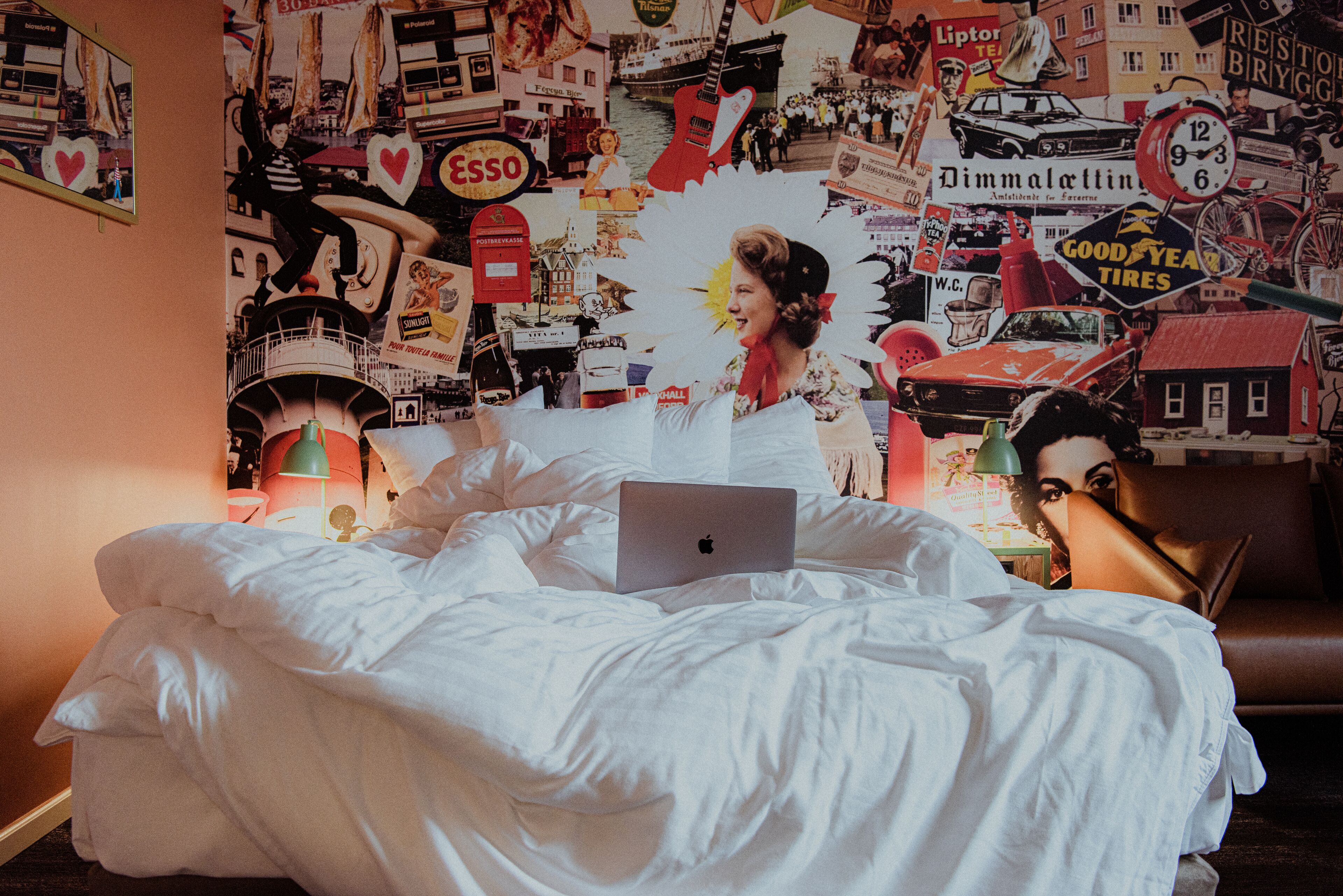 Un dormitorio con una cama, una laptop y una pared decorada con varios carteles y anuncios.
