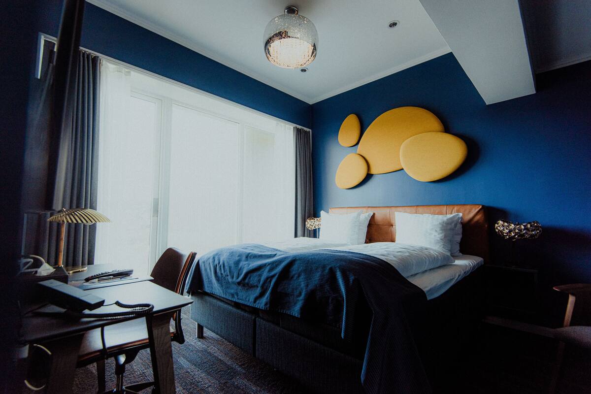 Une chambre d’hôtel avec un grand lit, un bureau et une chaise. Il y a une fenêtre avec des rideaux, une lampe et un élément décoratif au mur.