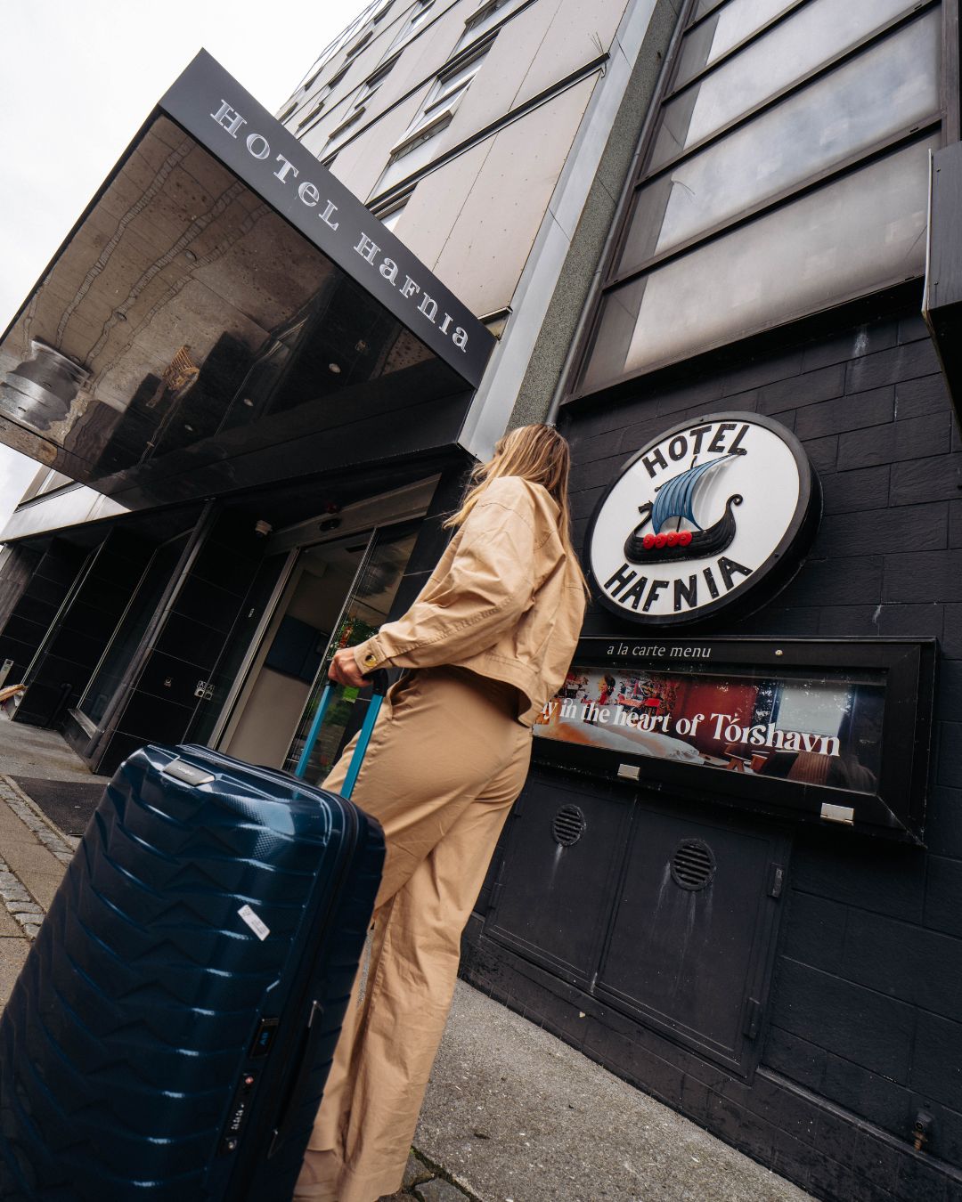 A person with a suitcase standing in front of a building with a sign that reads 'Hotel Hafnia.'