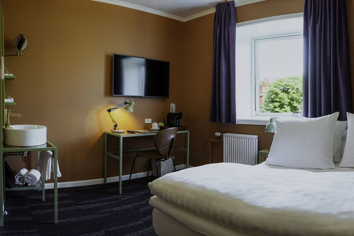 Une chambre d’hôtel avec un lit, un bureau avec une lampe, une télévision, une fenêtre avec des rideaux et un radiateur.
