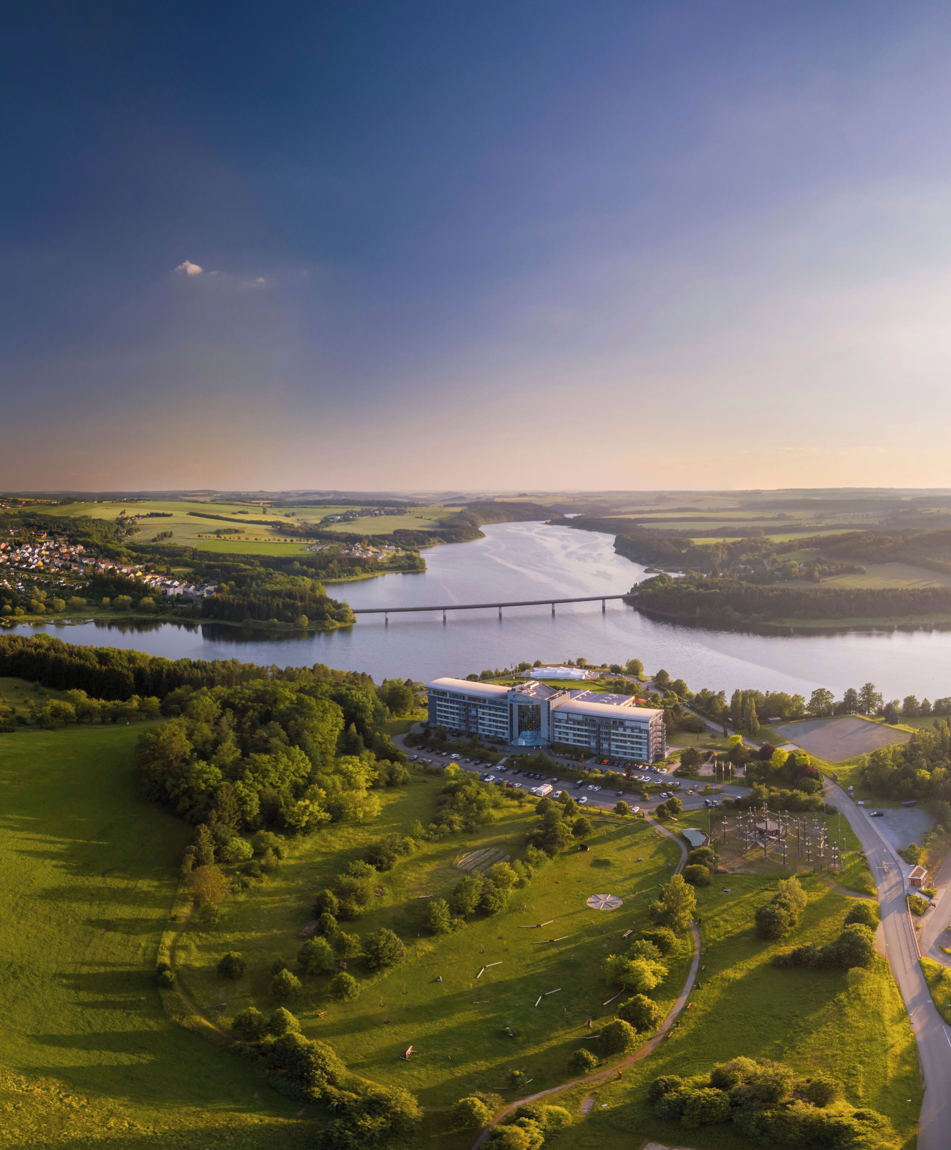 An aerial view of a hotel with a river in the background, surrounded by green fields and a bridge.