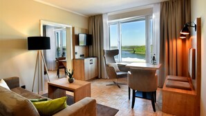 A modern hotel room with a wooden floor, a sofa, a dining table, a TV, and a view of a lake. - Bio-Seehotel Zeulenroda (Zeulenroda-Triebes)